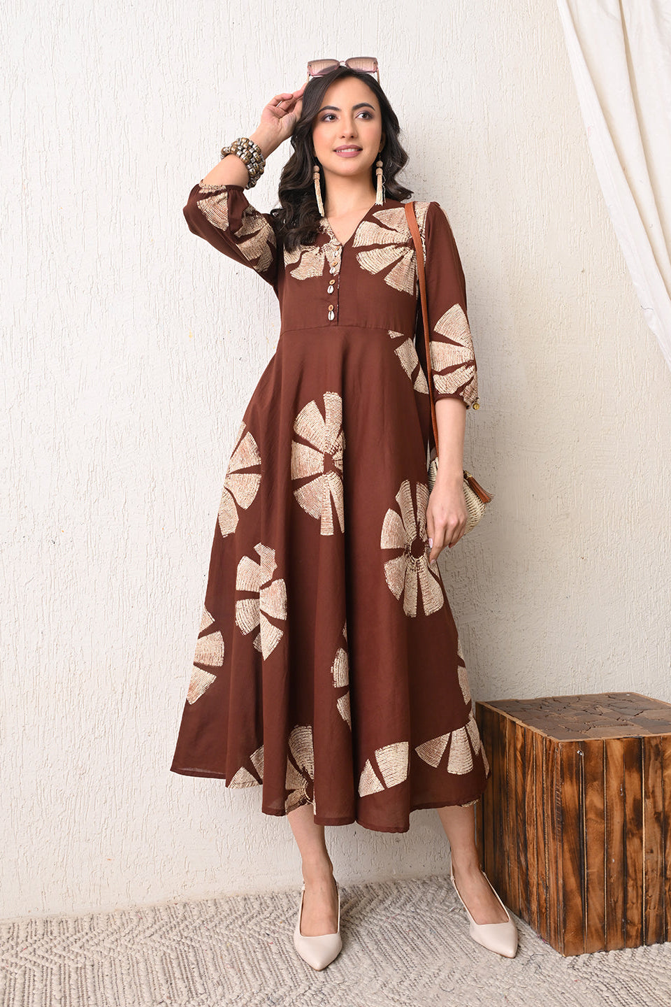 Woman wearing a brown floral dress from Desi Tassels standing against a white wall with a wooden box.