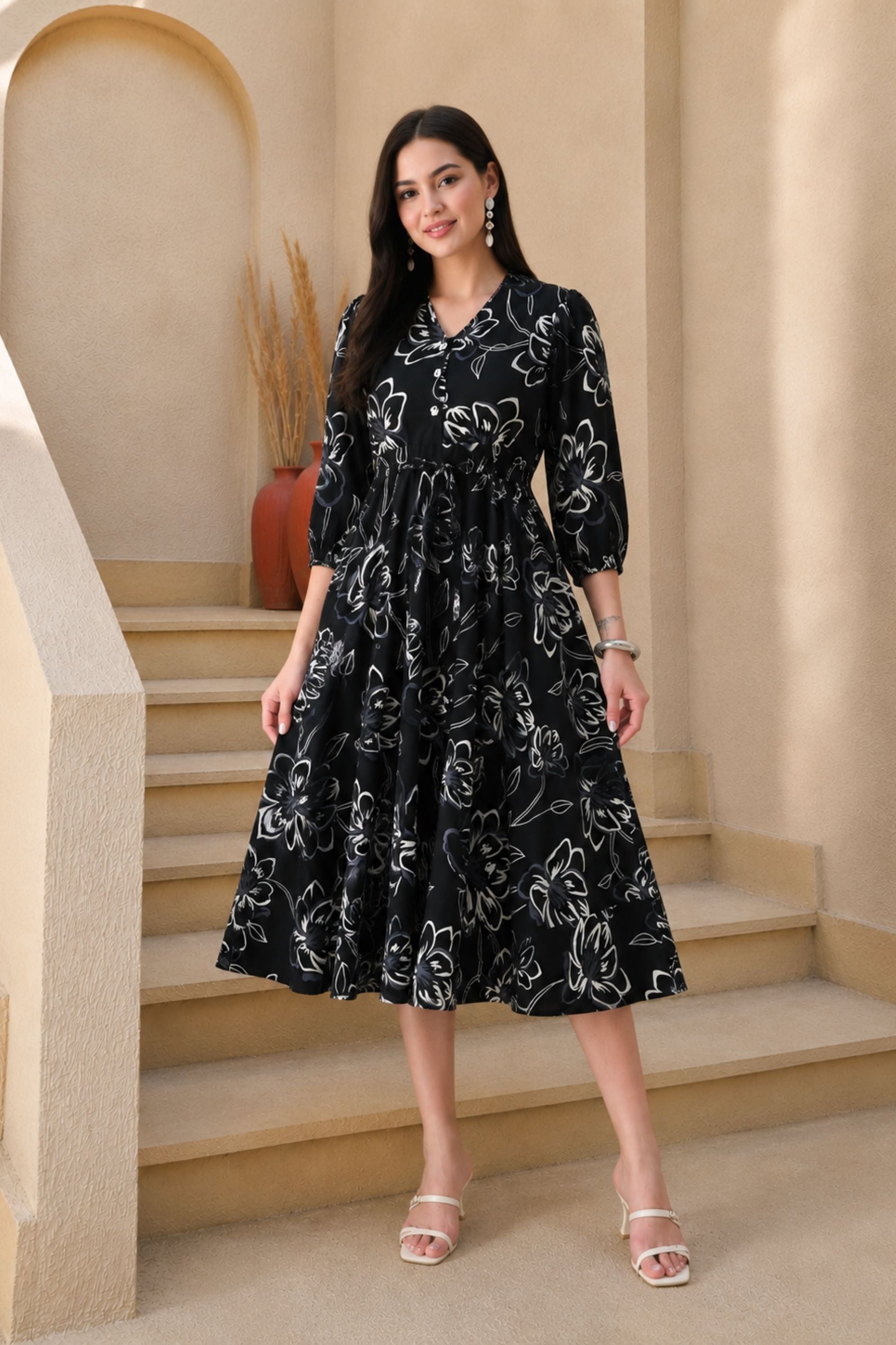 A woman stands on beige indoor stairs wearing a black midi dress with a white floral line pattern, three-quarter sleeves, and a V-neck. She is wearing clear heeled sandals and jewelry, with a warm neutral architectural background.