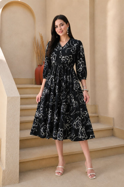 A woman stands on beige indoor stairs wearing a black midi dress with a white floral line pattern, three-quarter sleeves, and a V-neck. She is wearing clear heeled sandals and jewelry, with a warm neutral architectural background.