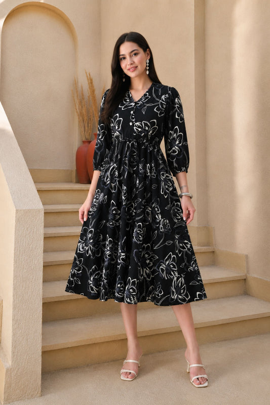 A woman stands on beige indoor stairs wearing a black midi dress with a white floral line pattern, three-quarter sleeves, and a V-neck. She is wearing clear heeled sandals and jewelry, with a warm neutral architectural background.
