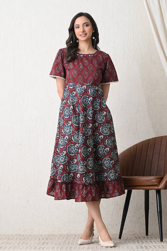 Woman wearing a patterned dress from Desi Tassels standing against a white wall with a chair in the background.