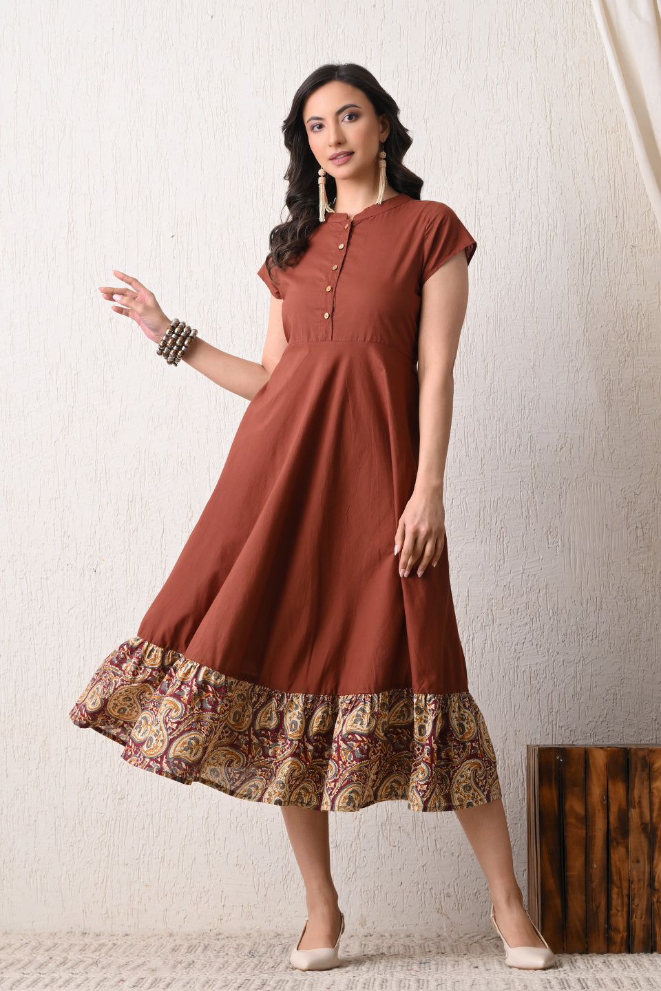 Woman wearing a brown dress with a patterned hem from Desi Tassels against a white wall.