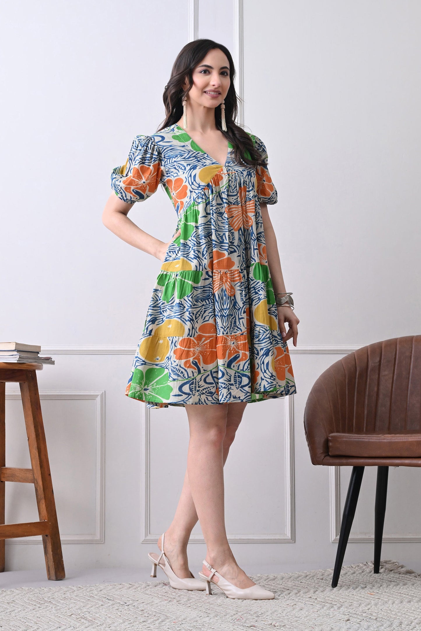 Woman wearing a colorful floral dress from Desi Tassels, standing in a room with a wooden chair and stool.