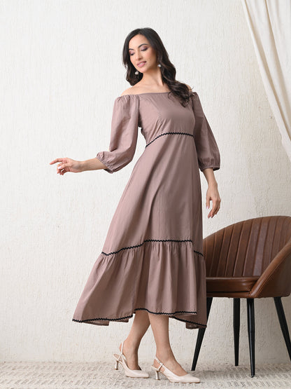 Woman wearing a brown dress from Desi Tassels with black trim standing in a room with a chair.