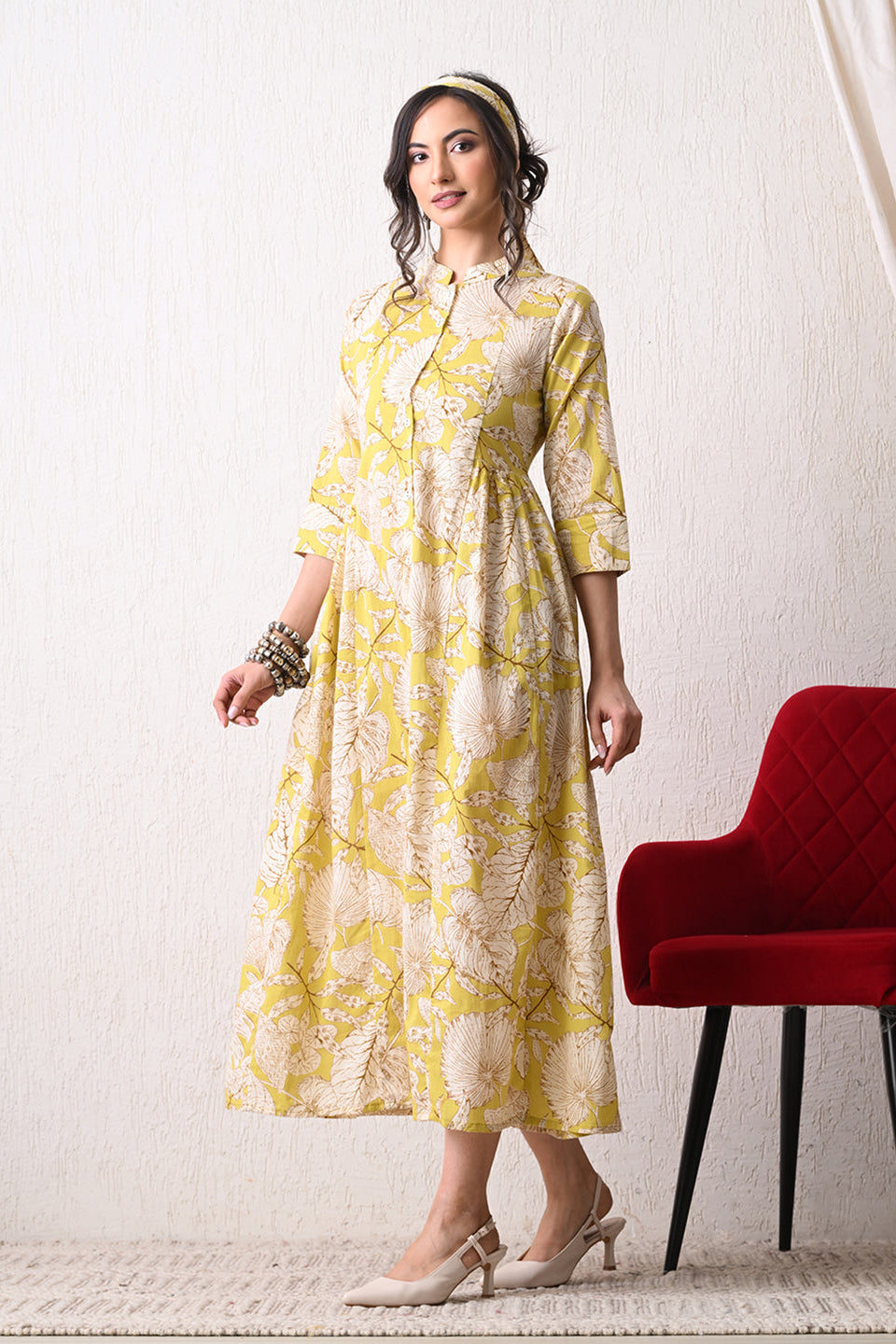 Woman wearing a yellow floral dress from Desi Tassels standing next to a red chair against a white wall.