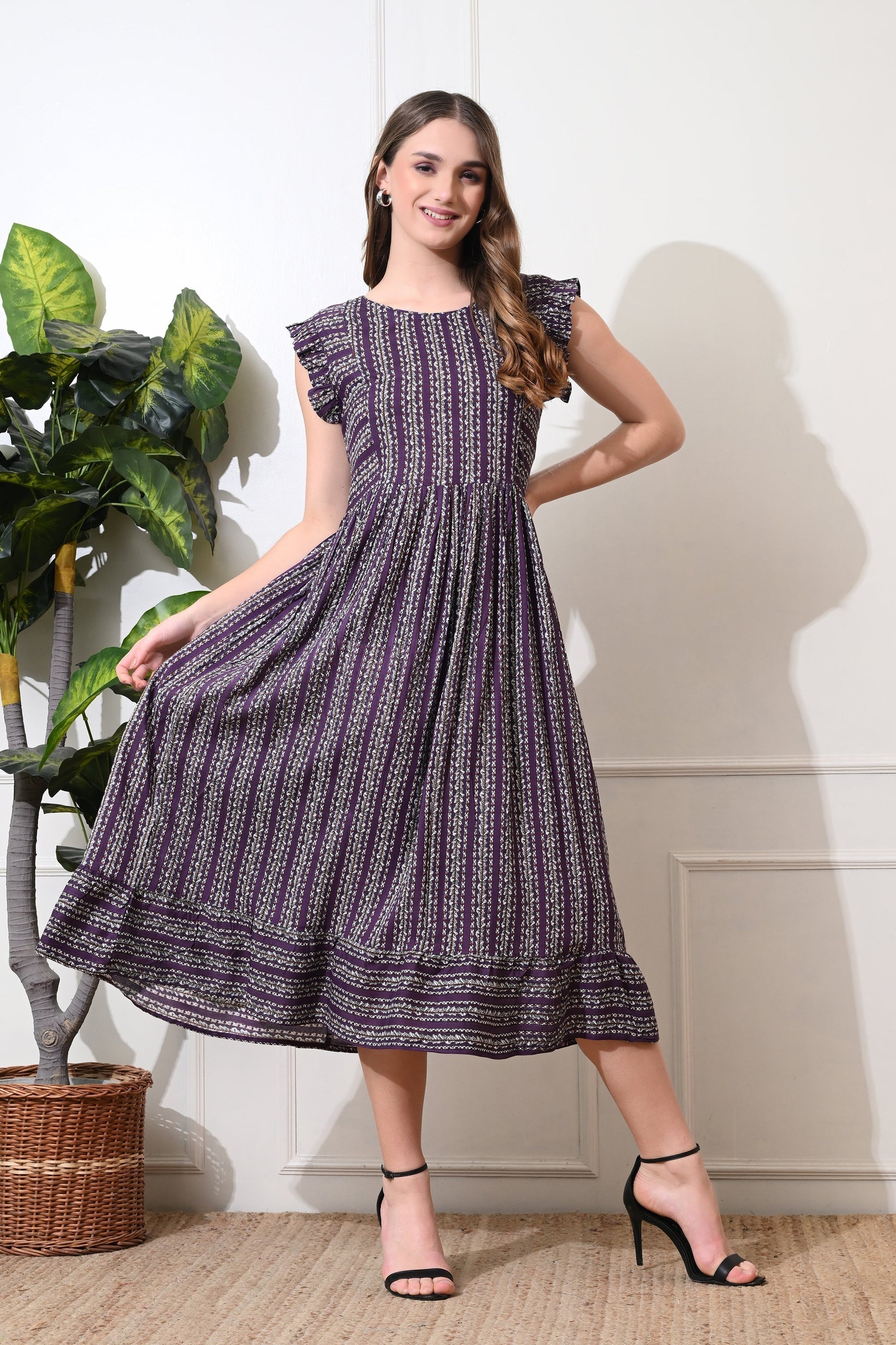 A woman standing in a room with a plant and a basket, wearing a green striped flared dress from Desi Tassels with a round neckline and sleeveless A-line silhouette.